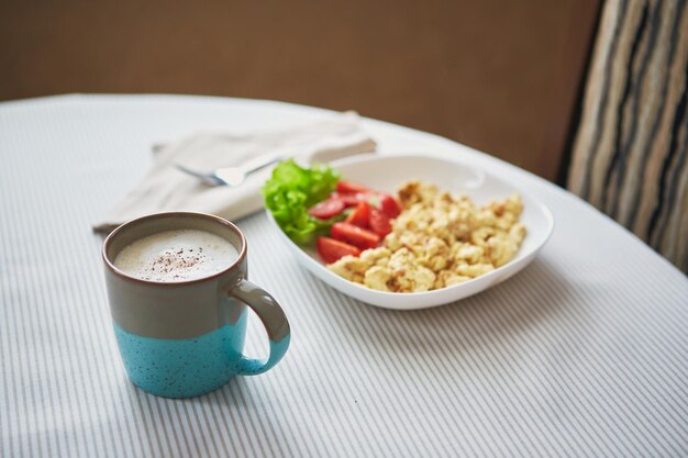 Café capuchino en taza y plato con desayuno de huevos revueltos y tomates en la mesa