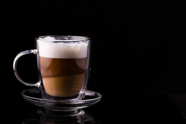 Café capuchino en una taza transparente sobre un fondo negro.