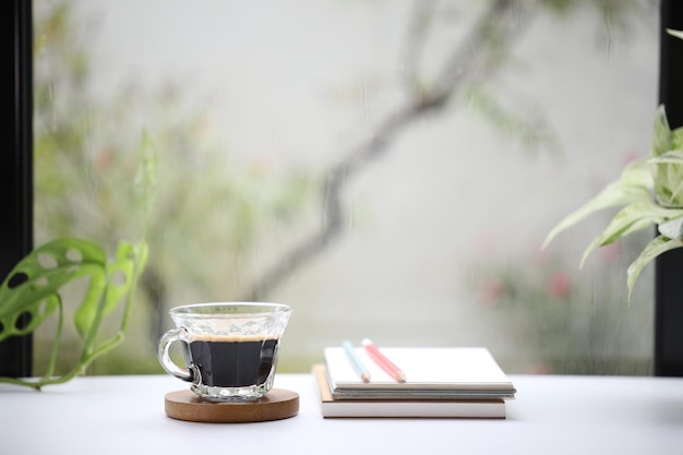 Café com crema em uma xícara de vidro e cadernos em mesa de madeira com plantas