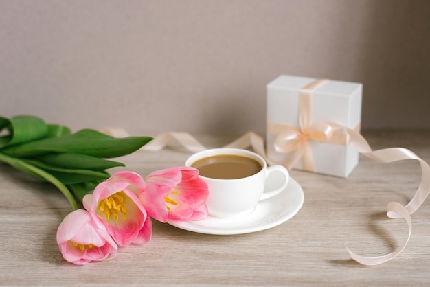 Café com leite em uma xícara de porcelana branca e pires um buquê de tulipas rosa primavera e um presente
