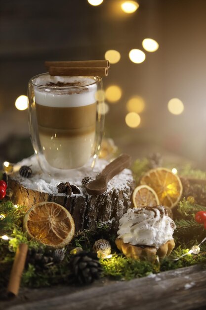 Café com leite festivo em uma tigela de vidro com tartelete no fundo escuro de madeira com laranja, canela e luzes