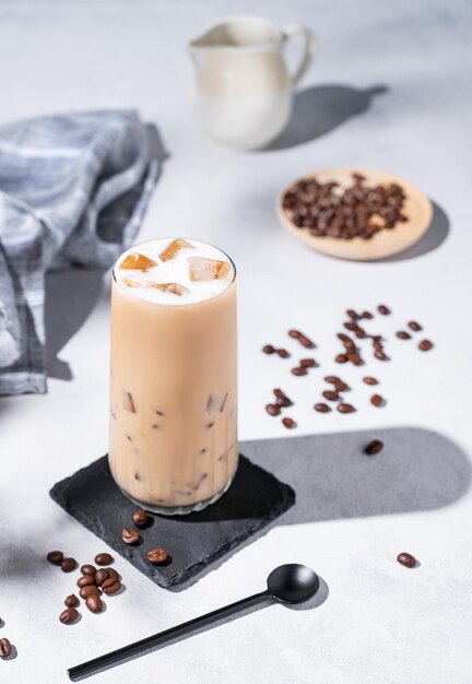 Café com leite gelado em um copo alto com leite em um fundo claro com grãos de café, jarro de leite e sombras matinais Conceito de refresco de verão