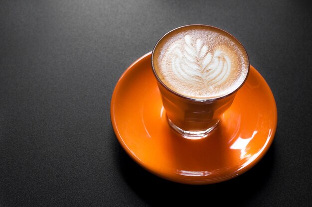 Café com leite quente em copo laranja com padrão floral em espuma em um fundo escuro