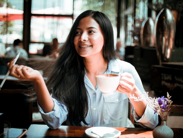 Café de consumición de la mujer asiática y trabajo con el ordenador portátil en café