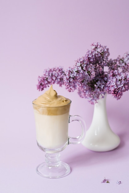 Café cremoso en taza de vidrio con flores púrpuras en jarrón blanco sobre fondo violeta claro