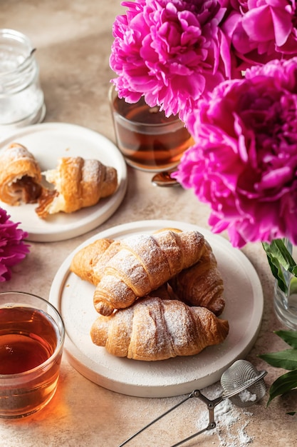 Café da manhã com dois pratos de açucareiro de chá croissants e flores de peônias em fundo marrom