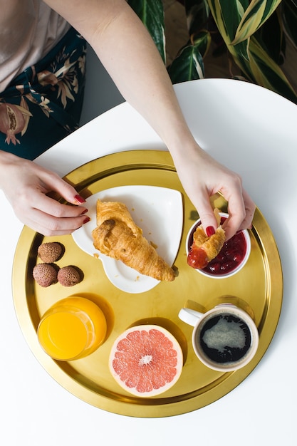 Café da manhã francês com croissant e café. As mãos das mulheres quebram o croissant. Café, geléia, croissant, suco de laranja, grapefruit, lichia.