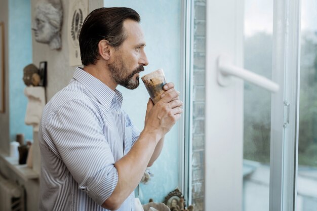 Café da manhã. homem barbudo em pé perto da janela tomando café da manhã antes de ir para o trabalho