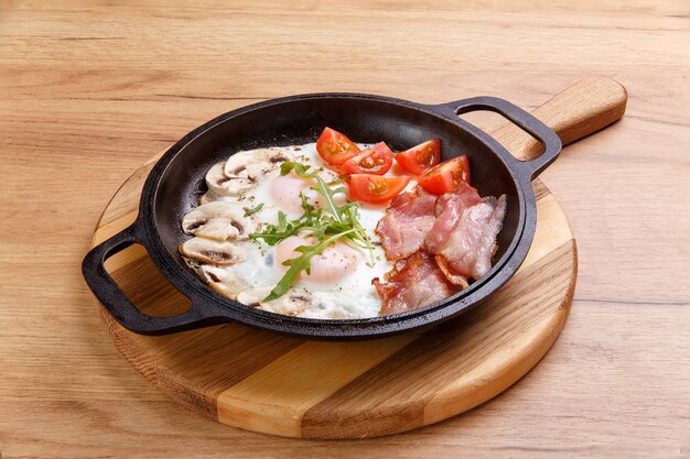 Café da manhã inglês na frigideira com ovos fritos, bacon, cogumelos e tomates em um suporte de madeira