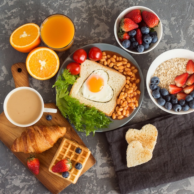 Café da manhã inglês: ovo frito, feijão e torradas em um prato com tomate cereja, salada, suco, frutas e croissant