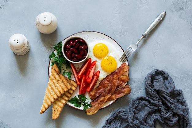 Café da manhã inglês tradicional com ovos fritos, bacon, feijão, torradas e tomates em concreto cinza