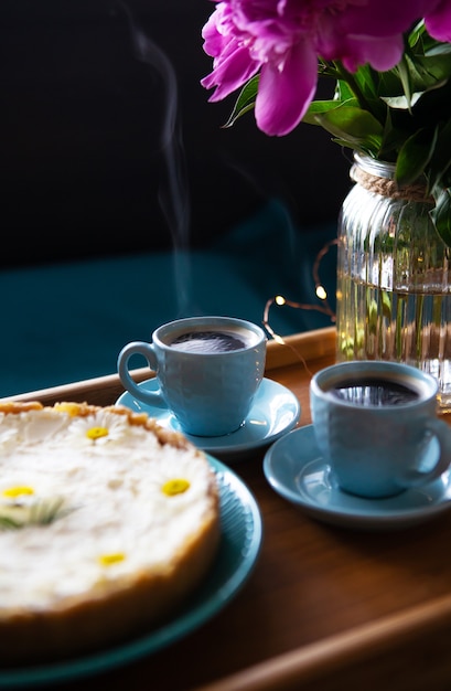 Café da manhã na cama. Lindas peônias, cheesecake delicioso e duas xícaras de café em uma bandeja de madeira.