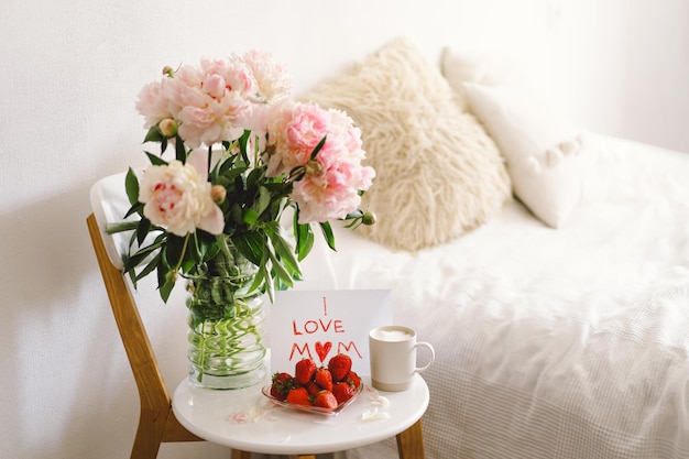 Café da manhã para o Dia das Mães Prato branco em forma de coração com morangos frescos, xícara de café e buquê de peônias com presente na cama Composição de natureza morta Feliz Dia das Mães