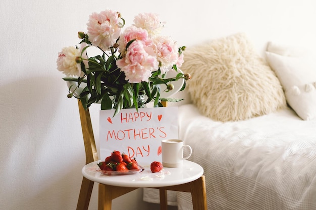 Café da manhã para o Dia das Mães Prato branco em forma de coração com morangos frescos, xícara de café e buquê de peônias com presente na cama Composição de natureza morta Feliz Dia das Mães