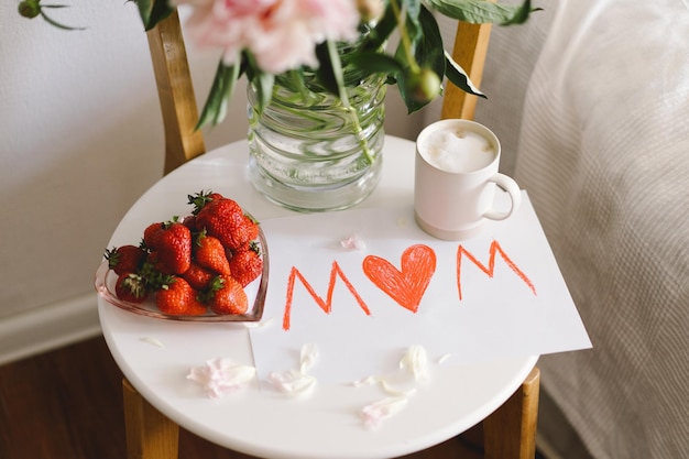 Café da manhã para o Dia das Mães Prato branco em forma de coração com morangos frescos, xícara de café e buquê de peônias com presente na cama Composição de natureza morta Feliz Dia das Mães