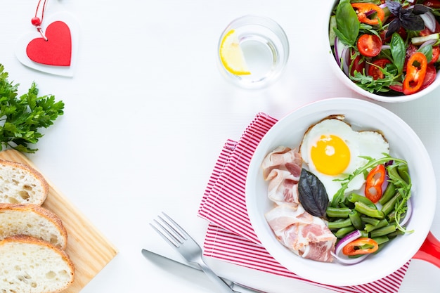 Café da manhã para o seu amado para o feriado: ovo em forma de coração, bacon, feijão verde em uma mesa branca. Foco seletivo. Vista do topo. Copie o espaço