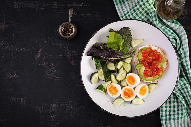 Café da manhã Sanduíche aberto saudável na torrada com abacate e caviar vermelho ovos cozidos salada de pepino no prato branco Comida de proteína saudável Vista superior