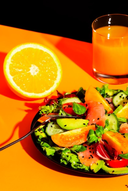 Foto café da manhã saudável com salada de salmão com sementes de gergelim abacate tomates ervas misturadas e suco de laranja natureza morta contemporânea fundo preto laranja luz dura e sombras duras