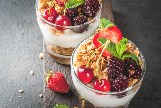 Café da manhã saudável. frutas e bagas de verão. iogurte grego caseiro com granola, amoras, morangos, cerejas e hortelã. na mesa de madeira e pedra preta, em copos.