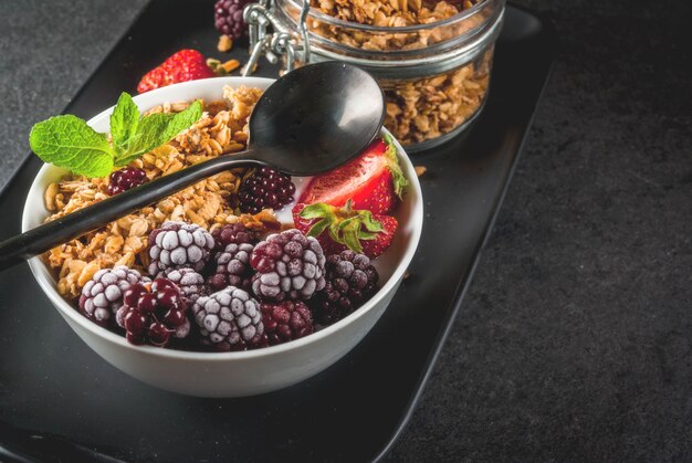 Foto café da manhã saudável. frutas e bagas de verão. iogurte grego caseiro com granola, amoras, morangos e hortelã. mesa de pedra preta, com os ingredientes.