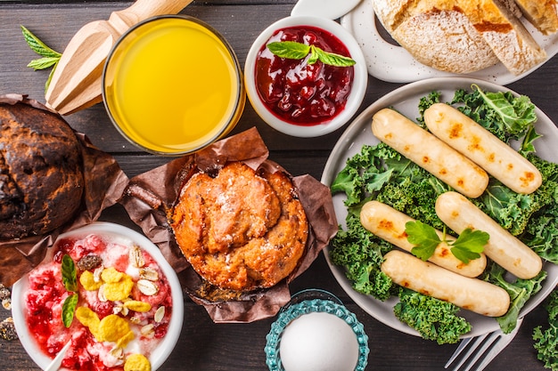 Café da manhã servido com bolinhos, salsichas grelhadas, suco, pão fresco e parfait em uma mesa de madeira escura.