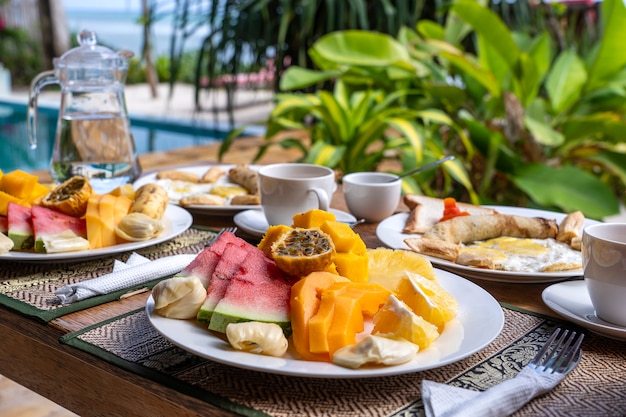 Café da manhã tropical de frutas, café e ovos mexidos e panqueca de banana para dois na praia, perto do mar