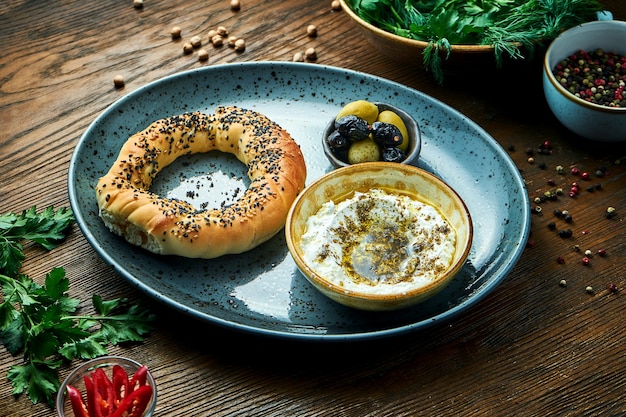 Café da manhã turco clássico - simit com mousse de queijo feta com azeite e especiarias, servido em prato azul com azeitonas. Mesa de madeira. Comida do restaurante. Simit - bagel com sementes de papoula ou sementes de gergelim
