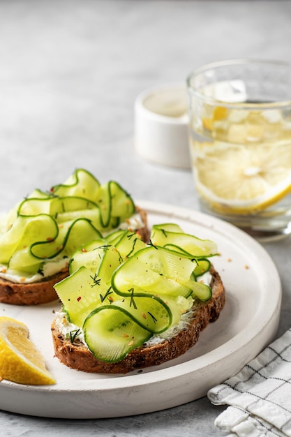 Café da manhã vegetariano saudável com sanduíches de pepino com queijo macio e água de limão em fundo cinza