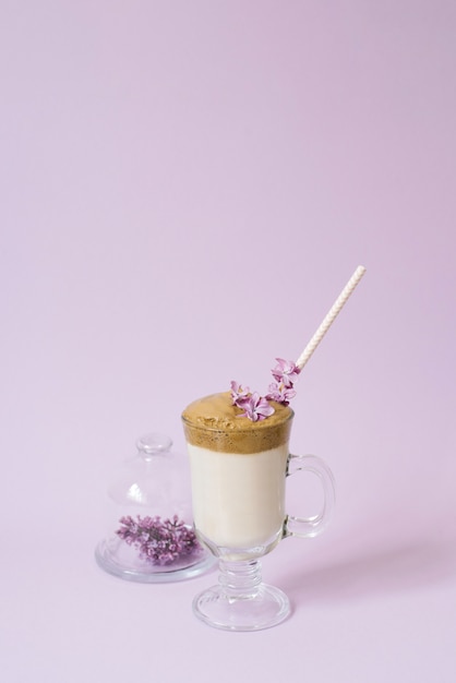 Café Dalgona en una taza transparente con un tubo de papel, un plato con flores lilas sobre lila