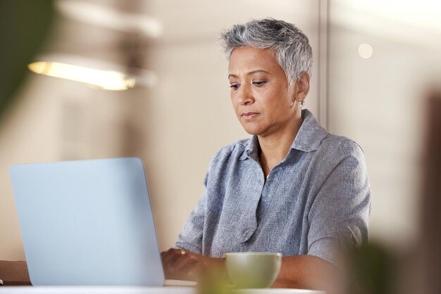 Café de escritório e mulher madura com ideia de digitação de laptop para negócios criativos de inicialização ou relatório na mesa Designer ou escritor autônomo sênior pensando e trabalhando em ideias no escritório moderno