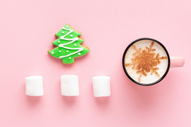 Café de Natal com biscoito e marshmallow em fundo rosa, layout plano. Pão de mel em forma de árvore de abeto e café com leite decorado com floco de neve.