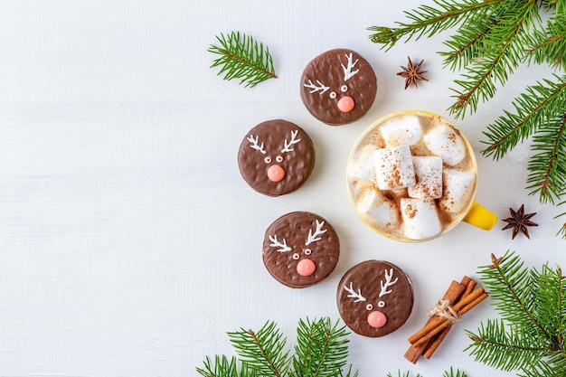 Café de Natal com marshmallows e biscoitos de chocolate com rostos de veado em fundo branco decorado com galhos de pinheiro.