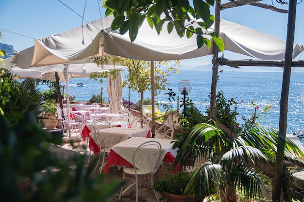 Café de verão na costa do mar Mesas com toalhas de mesa brancas sob um toldo do sol quente de grandes arbustos verdes Descanse em um dia quente de verão