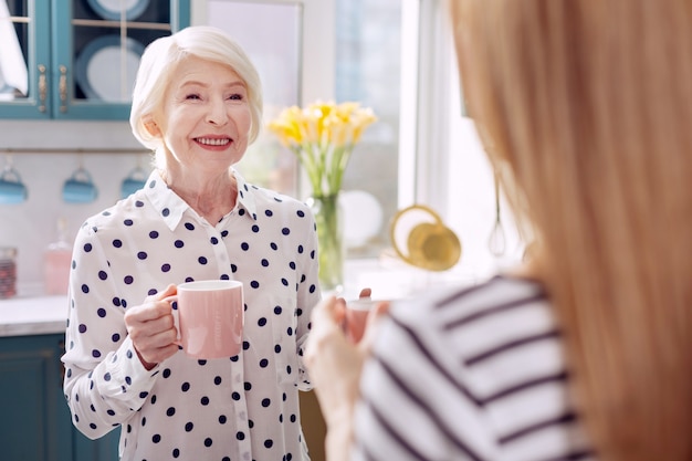 Café delicioso. o foco está em uma encantadora mulher sênior em pé na cozinha, tomando café com sua filha e conversando com ela