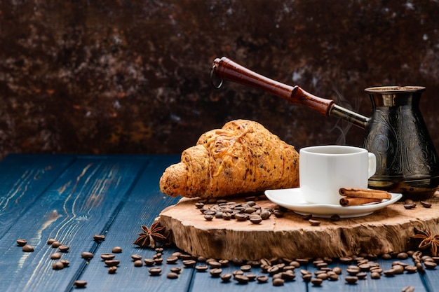 Café derrama em uma xícara sobre uma mesa de madeira azul com grãos de café e um croissant
