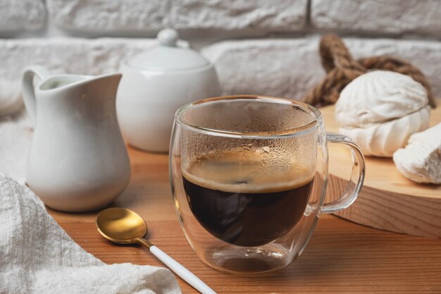 Café em uma caneca de vidro em uma mesa de madeira contra uma parede de tijolos brancos.