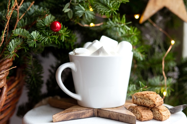 Café em uma xícara branca com marshmallows. Café festivo de manhã com biscoitos de amêndoa cantuccini italianos tradicionais. Uma xícara de café em um fundo de ramos de abeto verde em um suporte branco.