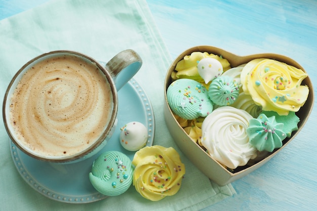 Café con espuma de leche en una taza azul y una caja en forma de corazón llena de merengues multicolores.