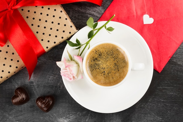 Café expresso em uma xícara branca, uma rosa, um presente com uma fita vermelha e chocolates em um fundo escuro. Vista do topo. Fundo de alimentos