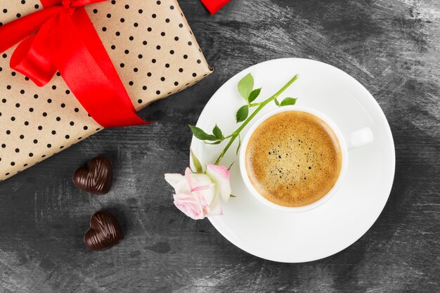 Café expresso em uma xícara branca, uma rosa, um presente com uma fita vermelha e chocolates em um fundo escuro. Vista do topo. Fundo de alimentos.