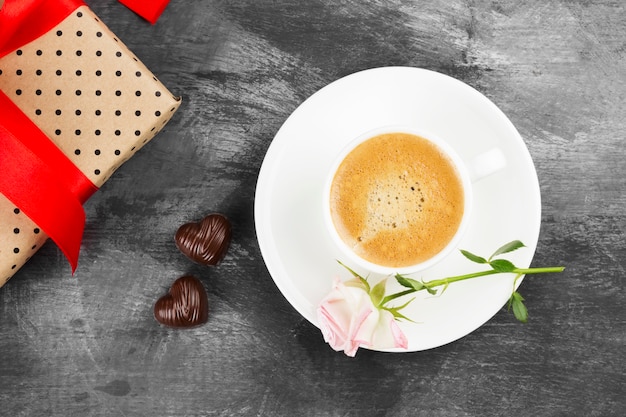 Café expresso em uma xícara branca, uma rosa, um presente com uma fita vermelha e chocolates em um fundo escuro. Vista do topo. Fundo de alimentos