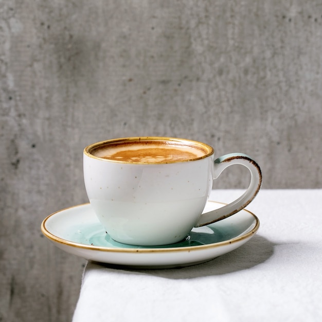 Café expresso preto com espuma em xícara de cerâmica branca com pires apoiado em toalha de mesa de algodão branco. Copie o espaço. Imagem quadrada