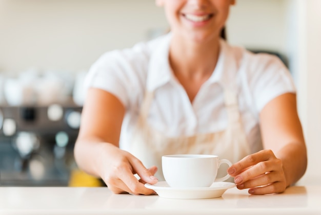 Café fresco para você. Close-up de uma bela jovem de avental servindo café e sorrindo em pé na cafeteria