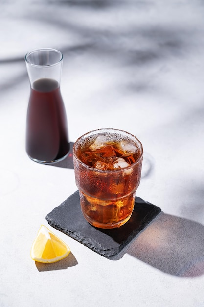 Café frío en un vaso con hielo en un fondo claro con botella de frutas de limón y sombras Concepto de artesanía de verano refrescante bebida casera