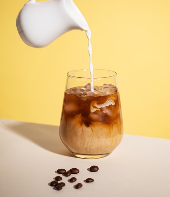 Café gelado com creme derramado em um copo com cubos de gelo e grãos em um fundo amarelo O conceito de uma bebida fria de verão