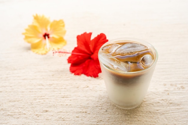 Café gelado com leite ao lado de hibisco na praia de areia.