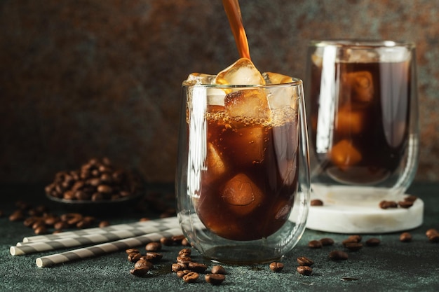 Café helado en un vaso alto con crema vertida sobre cubitos de hielo y frijoles en una mesa de hormigón oscura Bebida fría de verano con tubos sobre un fondo negro con espacio de copia