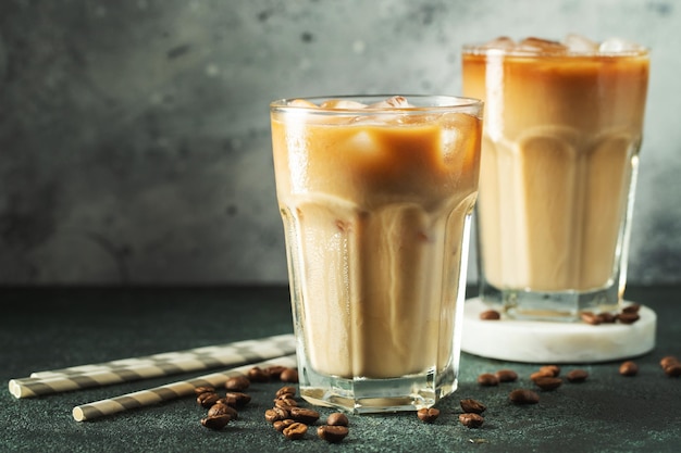 Café helado en un vaso alto con crema vertida sobre cubitos de hielo y frijoles en una mesa de hormigón oscura Bebida fría de verano con tubos sobre un fondo negro con espacio de copia