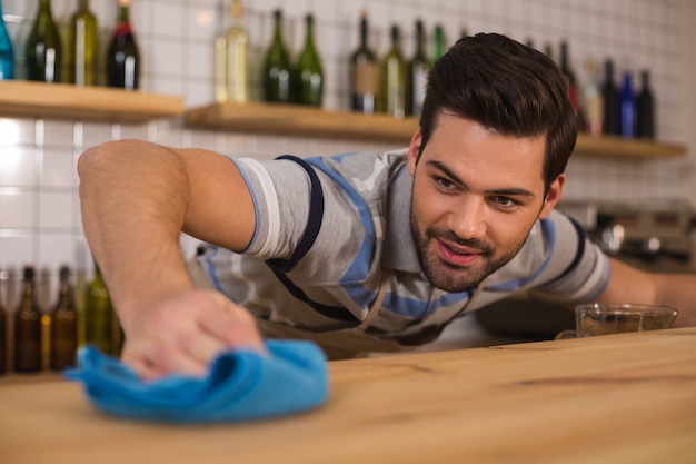 En el cafe. Hombre positivo agradable alegre sosteniendo el plumero y limpiando el mostrador mientras trabajaba en la cafetería