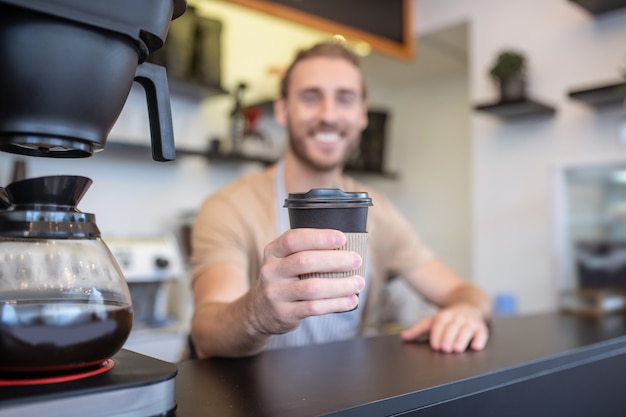 Café, humor. Copo de um delicioso café recém-feito na mão estendida de um sorridente jovem barista adulto atrás do balcão do bar